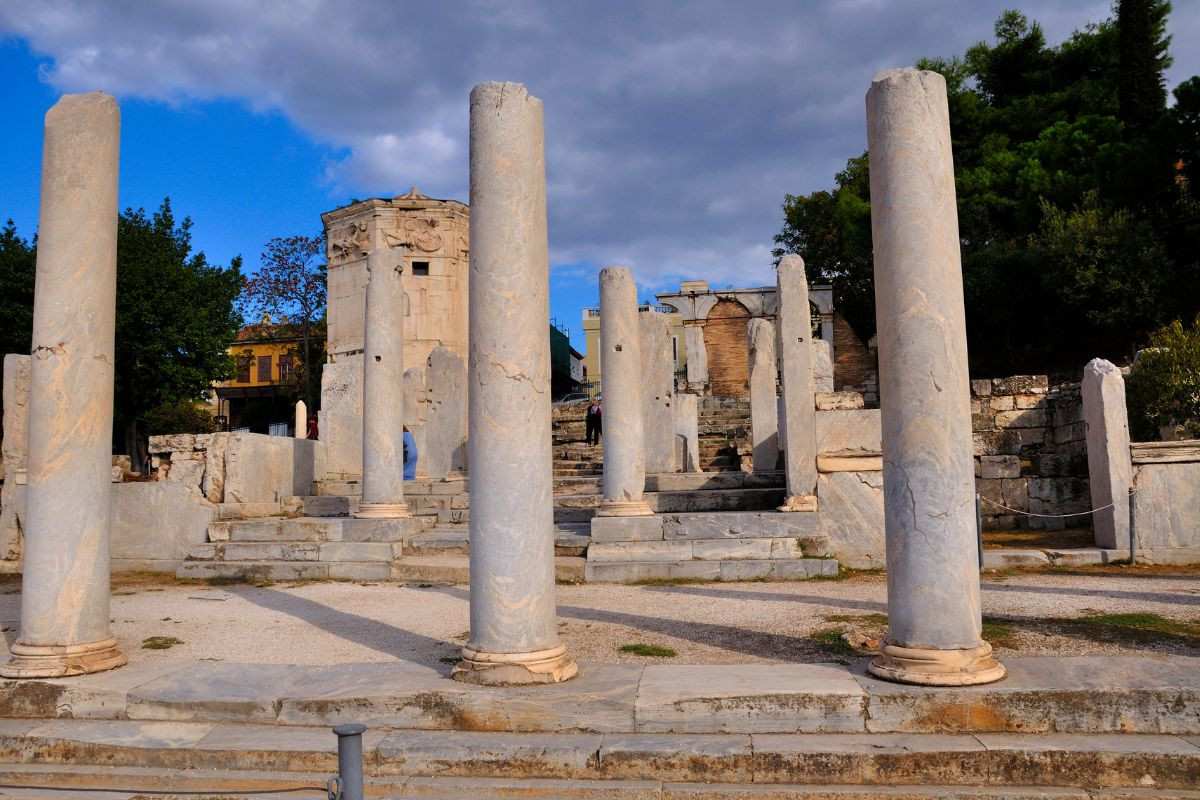 Ancient ruins of the Athens Agora, featuring weathered stone columns and the Tower of the Winds under a partly cloudy sky.